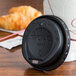 A Solo black hot cup lid on a coffee cup next to a croissant on a table.