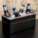 A black American Metalcraft rectangular hammered ice display with clear food pan holding bottles of beer on a table.