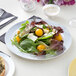 A Visions white plastic plate with silver lattice design holding a salad with tomatoes and greens.