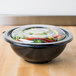 A Fineline clear plastic bowl with a lid on a wood surface filled with food.