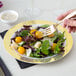 A hand holding a Visions gold lattice designed plastic fork over a salad on a Visions white plastic plate.