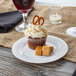 A Visions Wave white plastic plate with a cupcake with caramel and a pretzel on top next to a glass of wine.