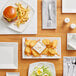 A wood table set with Acopa Bright White square porcelain plates of food including a hamburger and fries.