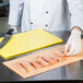 A person in a white coat and gloves uses a knife to cut raw meat on a yellow cutting board.