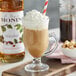 A glass of brown liquid with Monin Hazelnut syrup and whipped cream with a red and white straw.