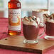 A glass mug of Monin Toasted Marshmallow hot chocolate with marshmallows and chocolate syrup on top.