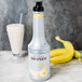 A white Monin bottle of banana puree next to a glass of banana juice.