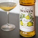 A bottle of Monin pear syrup next to a glass of yellow liquid with a Monin label.