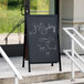 A black Aarco A-Frame chalkboard sign outside of a building with white writing.