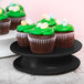 A black Elite Global Solutions melamine plate on a pedestal with cupcakes with green frosting and candy eggs.