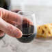 A hand holding a Fineline clear plastic wine goblet filled with red wine.