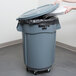 A person's hands using a dolly to lift a grey Rubbermaid trash can lid.