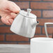 A hand holding a Vollrath Triennium stainless steel teapot over a white mug.