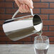 A hand pouring water from a Vollrath stainless steel pitcher into a glass.