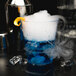 A clear WNA Comet plastic rocks glass filled with a blue drink and ice with smoke and a slice of orange on top.