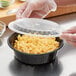A hand in a plastic glove putting a lid on a bowl of macaroni and cheese in a black plastic container.