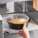 A hand placing a Choice round black plastic container of food in a microwave.