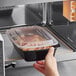A hand reaching out to a black Choice rectangular microwavable container filled with food.