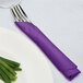 A fork and knife wrapped in an amethyst purple Creative Converting luncheon napkin on a white plate.
