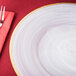 A white Charge It by Jay alabaster glass charger plate with a gold rim with a fork and plate on a red napkin.