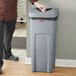 A man in a chef's uniform standing next to a Rubbermaid Untouchable gray square trash can.