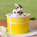 A yellow Choice paper cup filled with white and brown frozen yogurt topped with chocolate chips, with a white spoon.