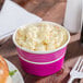 A pink paper food cup filled with macaroni and cheese next to a sandwich.