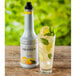 A bottle of Monin Yuzu Fruit Puree on a white background with a glass of lemonade and a glass of ice with mint leaves.
