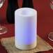 A white Sterno flameless cylindrical candle with a color changing feature on a table with burlap napkins and wine glasses.