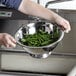 A person washing green beans in a Vollrath stainless steel colander on a counter.