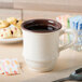 An ivory plastic coffee mug filled with coffee on a table with a spoon and pastries.