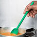 A hand holding a Mercer Culinary green high temperature mixing spoon over a bowl of food.