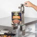 A hand opening a #10 can of beans on a stainless steel table with a Regency mobile can rack on the counter.