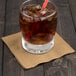 A glass with ice and a straw on a table with a Creative Converting Glittering Gold beverage napkin.