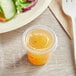 A plastic container of yellow sauce with a plastic lid on a table with a plate of salad and a cup of juice.