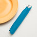 A turquoise blue Creative Converting paper dinner napkin wrapped around a fork on a table next to a plate.