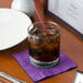 A glass of brown liquid with ice and a straw on a purple napkin.