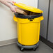 A person using a yellow Rubbermaid BRUTE trash can with lid to put a cardboard box inside.