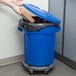 A person putting a box into a blue Rubbermaid BRUTE trash can with a lid.