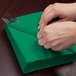 A person is wrapping a Creative Converting emerald green luncheon napkin around a fork and knife.
