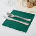 A knife and fork on a hunter green Creative Converting luncheon napkin next to a plate of food.