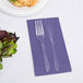 A fork and knife on a purple Creative Converting guest towel next to a plate of salad.