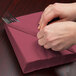 A person wrapping a burgundy Creative Converting luncheon napkin around a fork and knife.