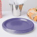 A stack of purple paper plates on a white background next to a silver bucket of sandwiches.
