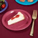 A Creative Converting burgundy paper plate with a slice of cheesecake topped with strawberry jam and a fork.
