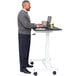 A man standing at a white Luxor stand up desk using a laptop.