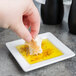 A hand holding a piece of bread over a Libbey Ultra Bright White wide rim square porcelain plate filled with olive oil.