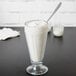 A glass of Ghirardelli Sweet Ground White Chocolate milkshake with a straw and a spoon on a table.