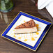 A slice of cheesecake on a Fineline white plastic plate with blue rim and gold bands.