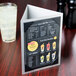 A Menu Solutions Alumitique triple view aluminum menu display on a table with a glass of juice.